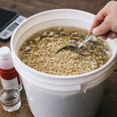 A hand holding a spoon stirs soaked barley grains in a white bucket filled with water, with small bottles beside it on a wooden table.