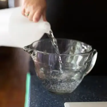 A hand pours clear liquid from a white plastic jug into a glass measuring bowl on a dark kitchen countertop.