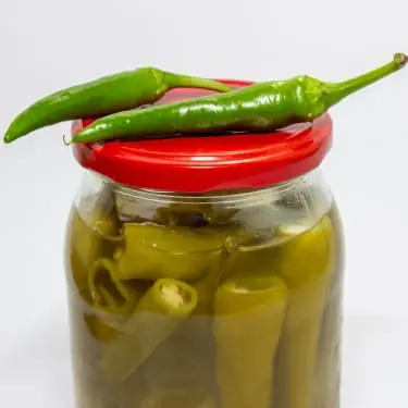 "A glass jar with a red lid filled with pickled green chili peppers in brine, with two fresh green chili peppers resting on top of the lid, against a plain white background.