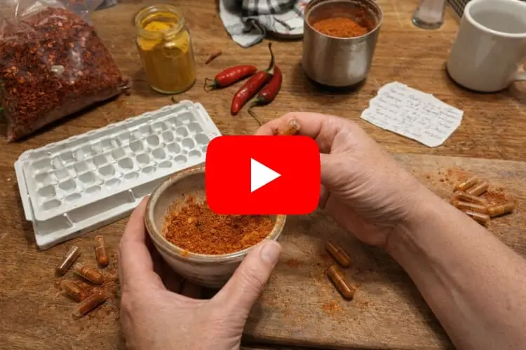 Two hands filling a clear gelatin capsule with orange cayenne pepper powder from a small ceramic bowl on a wooden table, surrounded by scattered filled capsules, a white capsule filling tray, a bag of red pepper flakes, a jar of turmeric, fresh red chili peppers, a handwritten recipe note, and a coffee mug