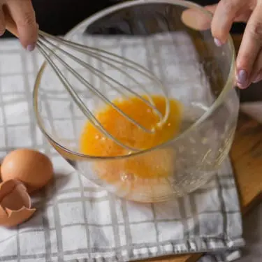 Two hands whisking eggs in a clear glass bowl on a checkered kitchen towel, with cracked eggshells and a wooden cutting board nearby