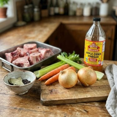 Bone broth ingredients on a rustic wooden cutting board raw beef bones in a metal tray, onions, carrots, celery, a small bowl of peppercorns and bay leaves, and a bottle of Bragg organic apple cider vinegar in a farmhouse-style kitchen