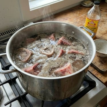 Beef bones simmering in a large stainless steel pot of water on a gas stove, with a bottle of apple cider vinegar and a small bowl in the background.