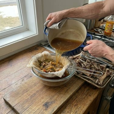 Hands pouring bone broth through a cheesecloth-lined strainer into a glass bowl on a wooden cutting board, with a tray of cooked bones and a gas stove in the background.