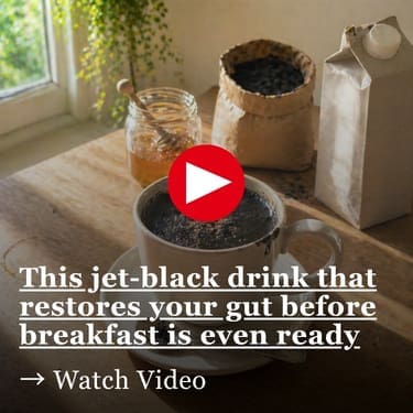 A sunlit kitchen table holds a white ceramic mug on a saucer, filled with an unusual jet-black drink topped with a light dusting of brown spice. A spoon with a small dark smear rests on the saucer, while a jar of honey, a crumpled paper bag of black powder, and an oat milk carton sit softly out of focus behind it. Warm morning light from a nearby window casts long shadows across the worn wooden surface, highlighting a faint ring stain and scattered crumbs.