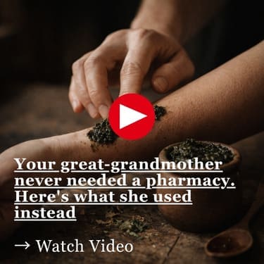 A hand applying a dark crushed herbal mixture onto the inner forearm, with a small clay pot filled with the same herbs and scattered dried leaves on a worn wooden surface beside a wooden spoon, in warm dim lighting.