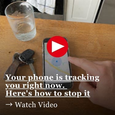 A top-down view of a black smartphone on a scratched wooden kitchen table, showing a map with a blue location dot, as a hand reaches in to tap the screen. A glass of water and a set of keys sit nearby, with a softly blurred kitchen in the background.