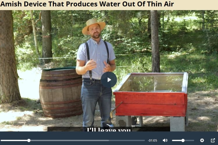 Video screenshot showing a man in a straw hat, light blue shirt, suspenders, and jeans standing outdoors beside a red glass-topped box on cinder blocks and a wooden barrel. Trees and grass fill the background, and the video title at the top reads, “Amish Device That Produces Water Out Of Thin Air.”