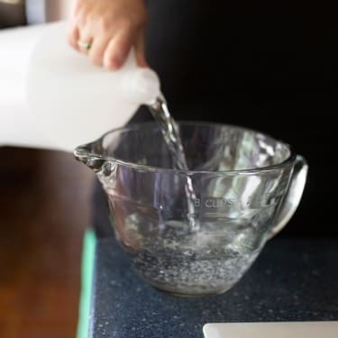 A hand pours clear liquid from a white plastic jug into a glass measuring bowl on a dark kitchen countertop.