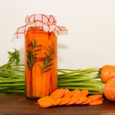 "A glass jar filled with pickled carrot sticks and a sprig of rosemary, topped with a red and white checkered cloth. Sliced carrot rounds, fresh whole carrots with green tops, and small orange onions are arranged on a wooden surface beside the jar, against a plain light background.