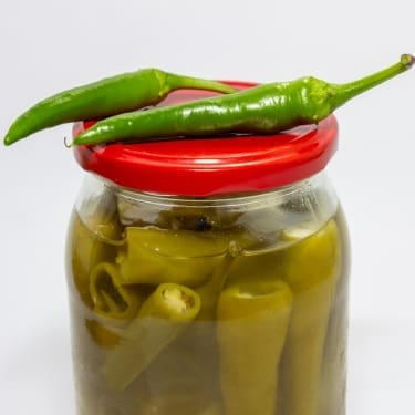 "A glass jar with a red lid filled with pickled green chili peppers in brine, with two fresh green chili peppers resting on top of the lid, against a plain white background.