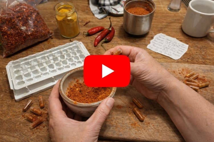 Two hands filling a clear gelatin capsule with orange cayenne pepper powder from a small ceramic bowl on a wooden table, surrounded by scattered filled capsules, a white capsule filling tray, a bag of red pepper flakes, a jar of turmeric, fresh red chili peppers, a handwritten recipe note, and a coffee mug