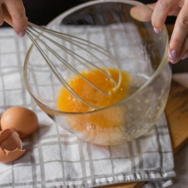 Two hands whisking eggs in a clear glass bowl on a checkered kitchen towel, with cracked eggshells and a wooden cutting board nearby