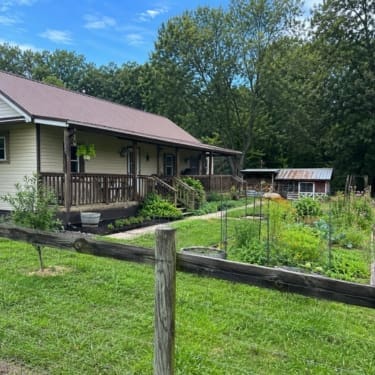 A sunny rural homestead in Tennessee with a single-story farmhouse, covered porch, fenced garden, grassy yard, and small outbuilding surrounded by tall green trees.