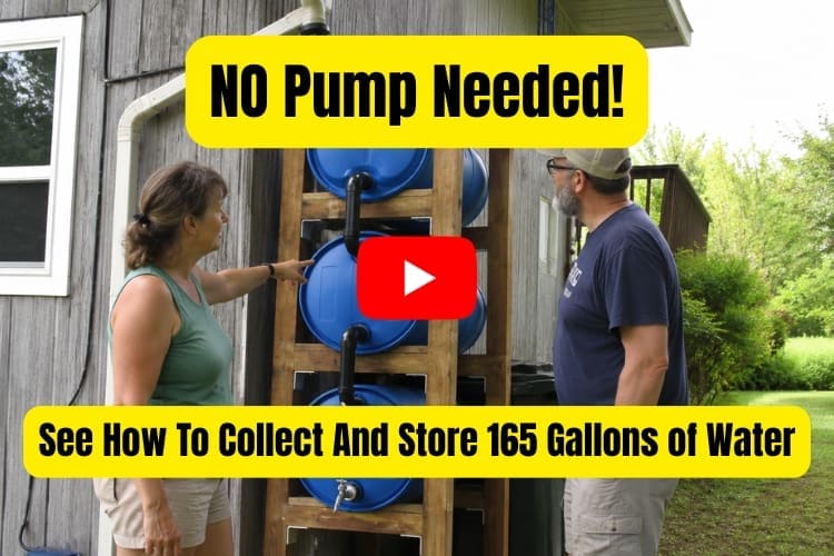 Two adults stand outside beside a homemade vertical rainwater harvesting system made from three stacked blue barrels in a wooden frame. Black pipes connect the barrels to a roof gutter, and a faucet is attached near the bottom barrel. One person points at the system while the other looks on, showing how the setup stores and gravity-pressurizes rainwater for garden use or filtering.