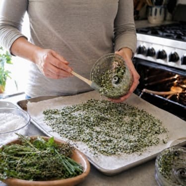 A person in a gray long-sleeve shirt scrapes a green herb-and-coarse-salt mixture from a food processor onto a parchment-lined baking sheet, with fresh herbs, coarse salt, and a home kitchen stove nearby.
